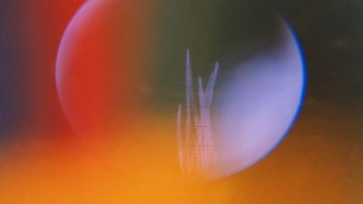 Film still from 'The Spheroids' (2025) by Jon Beaton. A blueish sphere is surrounded by red and orange light against a dark background.